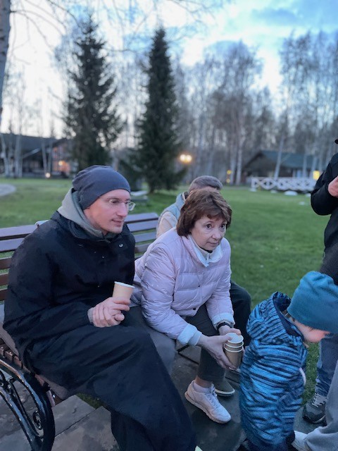 Мужчина в шапке и пожилая женщина в светлой куртке сидят на скамейке в парке с бумажными стаканами, рядом ребёнок